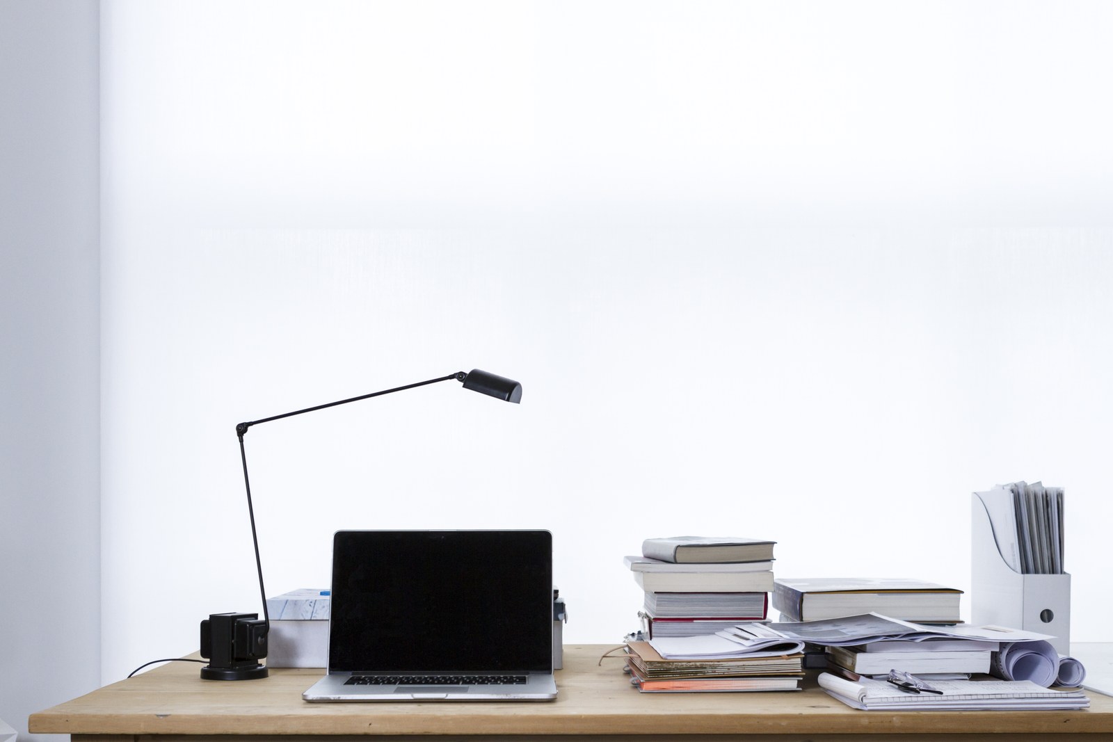 Mesa de trabajo con un portátil de pantalla negra, lámpara y pilas de documentos.